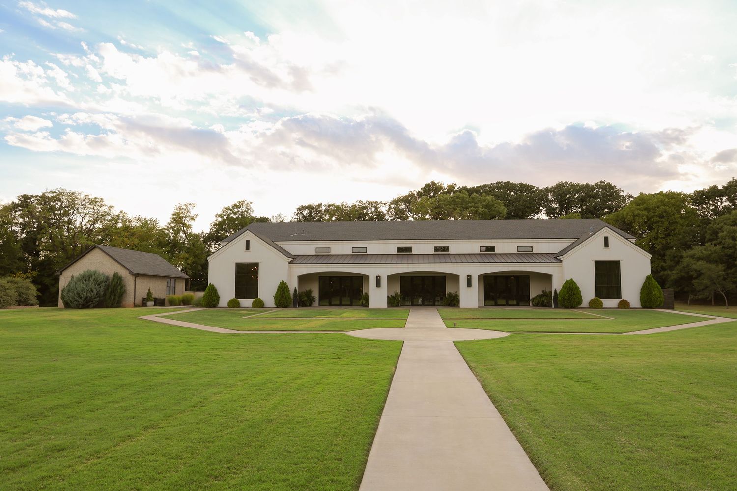 White barn venue with manicured lawn and curved pathway under a dramatic sky surrounded by lush trees.