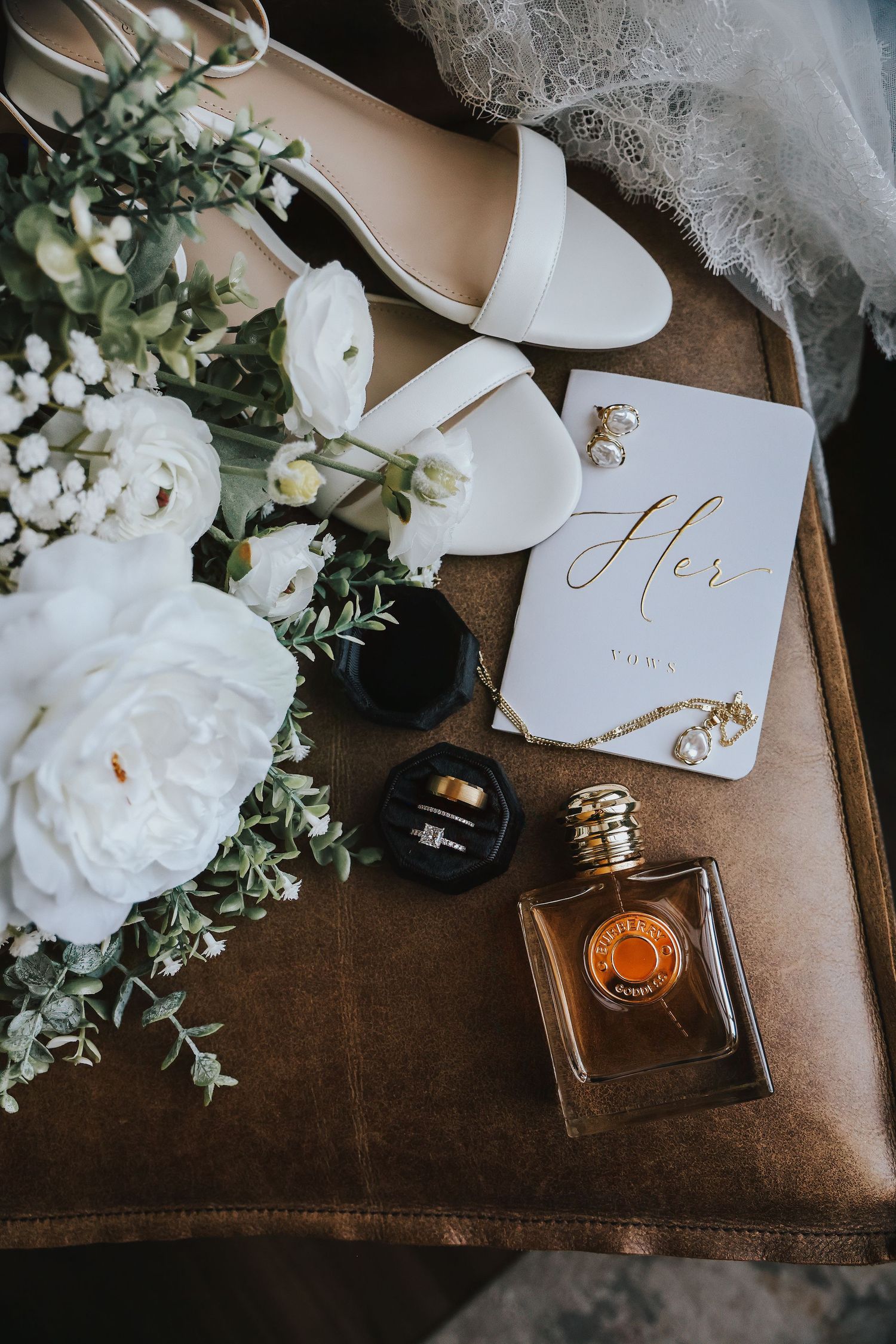 Bridal flat lay with white heels, floral bouquet, perfume bottle, ring box, pearl earrings, and a personalized wedding vow book.