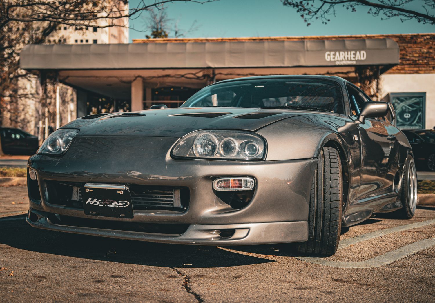 Silver Toyota Supra MK4 parked in front of a Gearhead shop, showcasing its iconic wide-body styling.