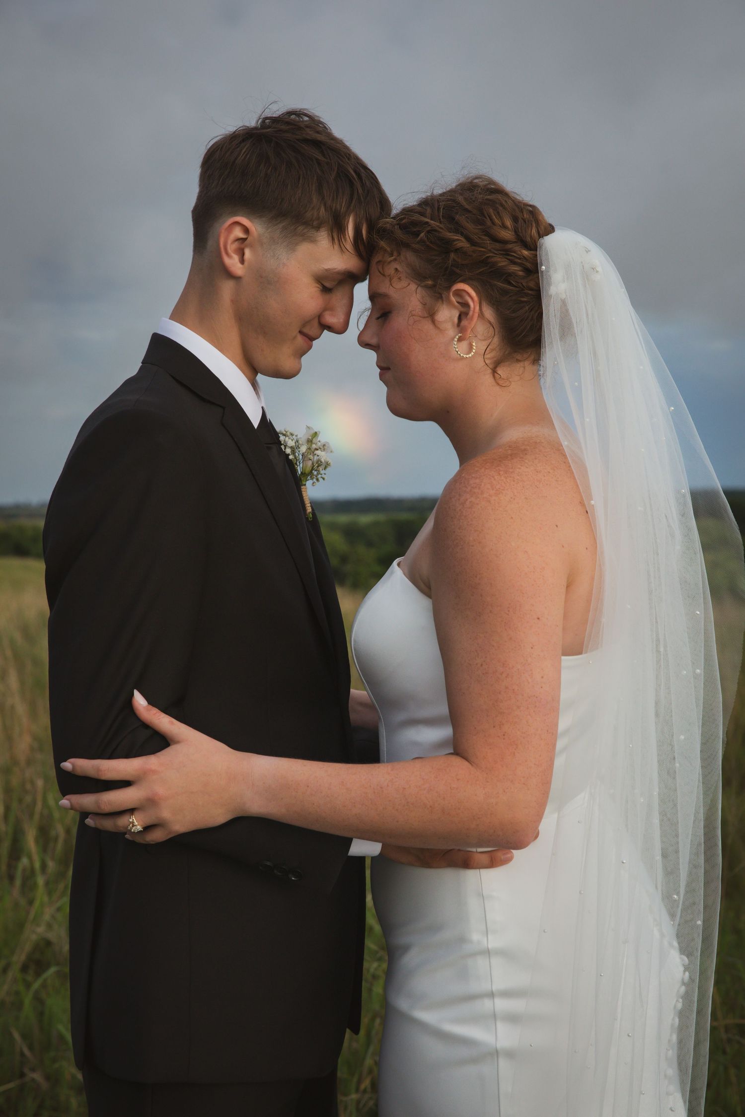 Bride and groom touching foreheads in a tender moment outdoors, with a rainbow visible in the cloudy sky behind them.