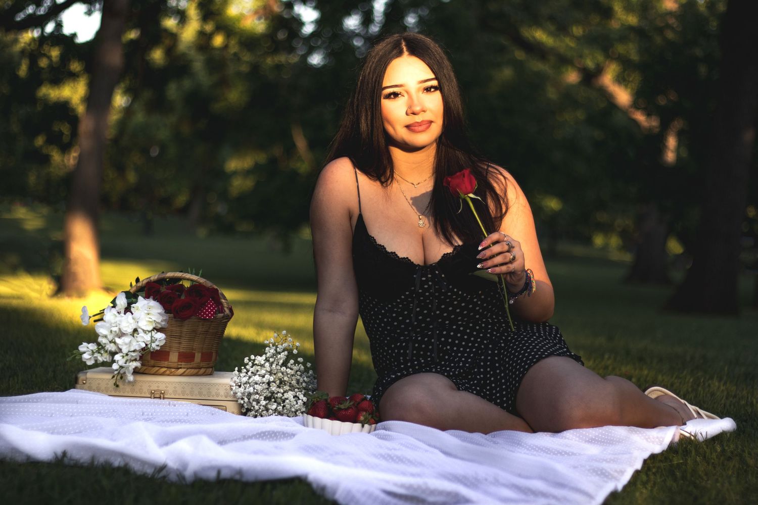 Young woman in black polka dot dress sits on white blanket in park, holding rose near flower basket at golden hour.
