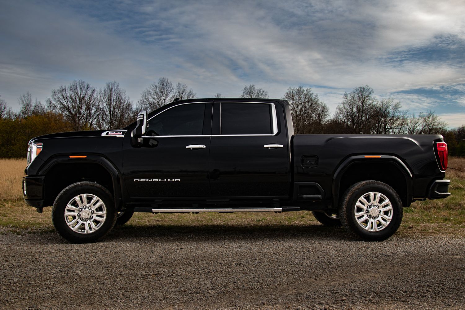 Black GMC Sierra HD crew cab pickup truck parked on gravel under dramatic cloudy sky, side profile view.