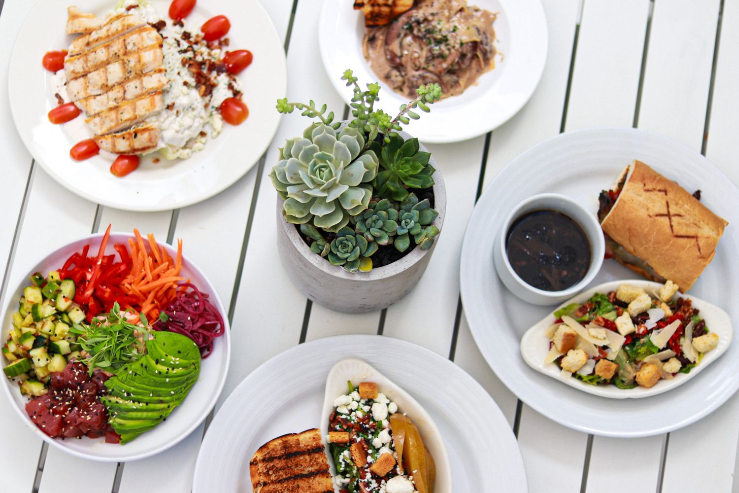 Overhead view of various plated dishes and drinks on a white wooden table setting.