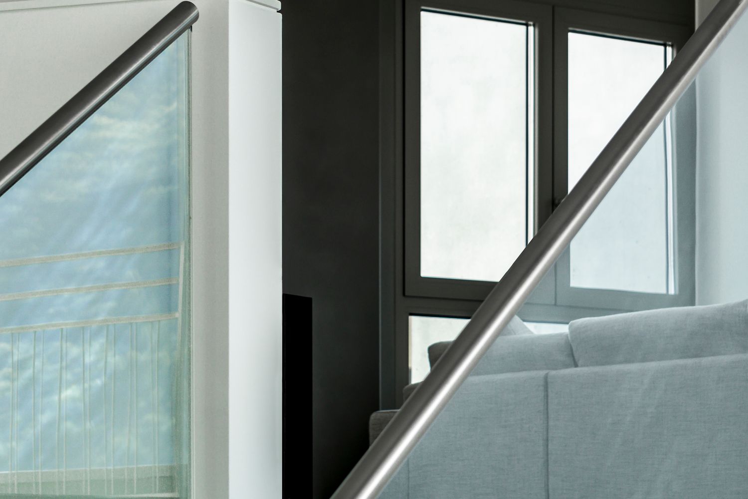 Dettaglio di interni moderni con corrimano metallico, vetro e divano chiaro, in un ambiente luminoso con grandi finestre.