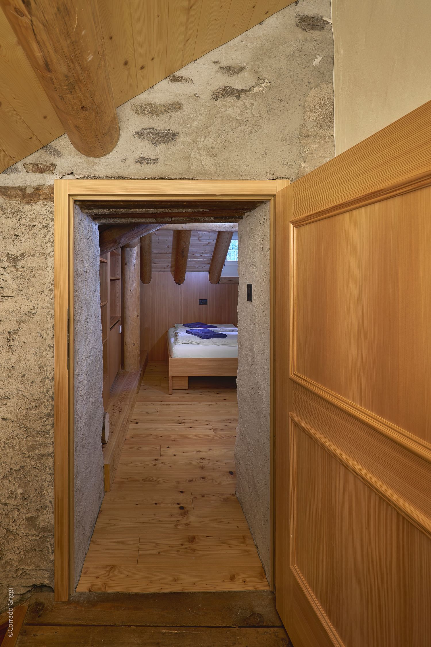 ingresso in pietra verso una camera mansardata in legno, con travi a vista e letto essenziale: fusione di elementi rustici e interior design