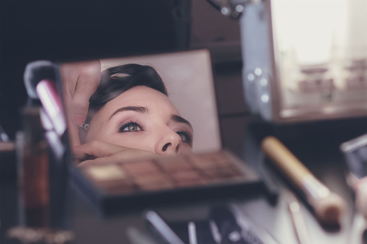 «Sguardo riflesso di una giovane donna in un piccolo specchio da trucco, con cosmetici sfocati attorno che creano un’atmosfera intima.»