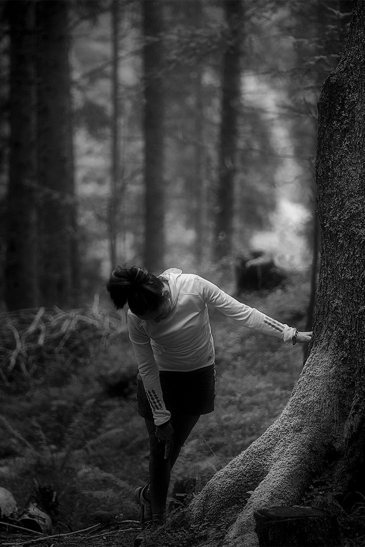 Figura femminile in ascolto accanto a un albero. In bianco e nero, il corpo diventa ponte tra natura e interiorità.