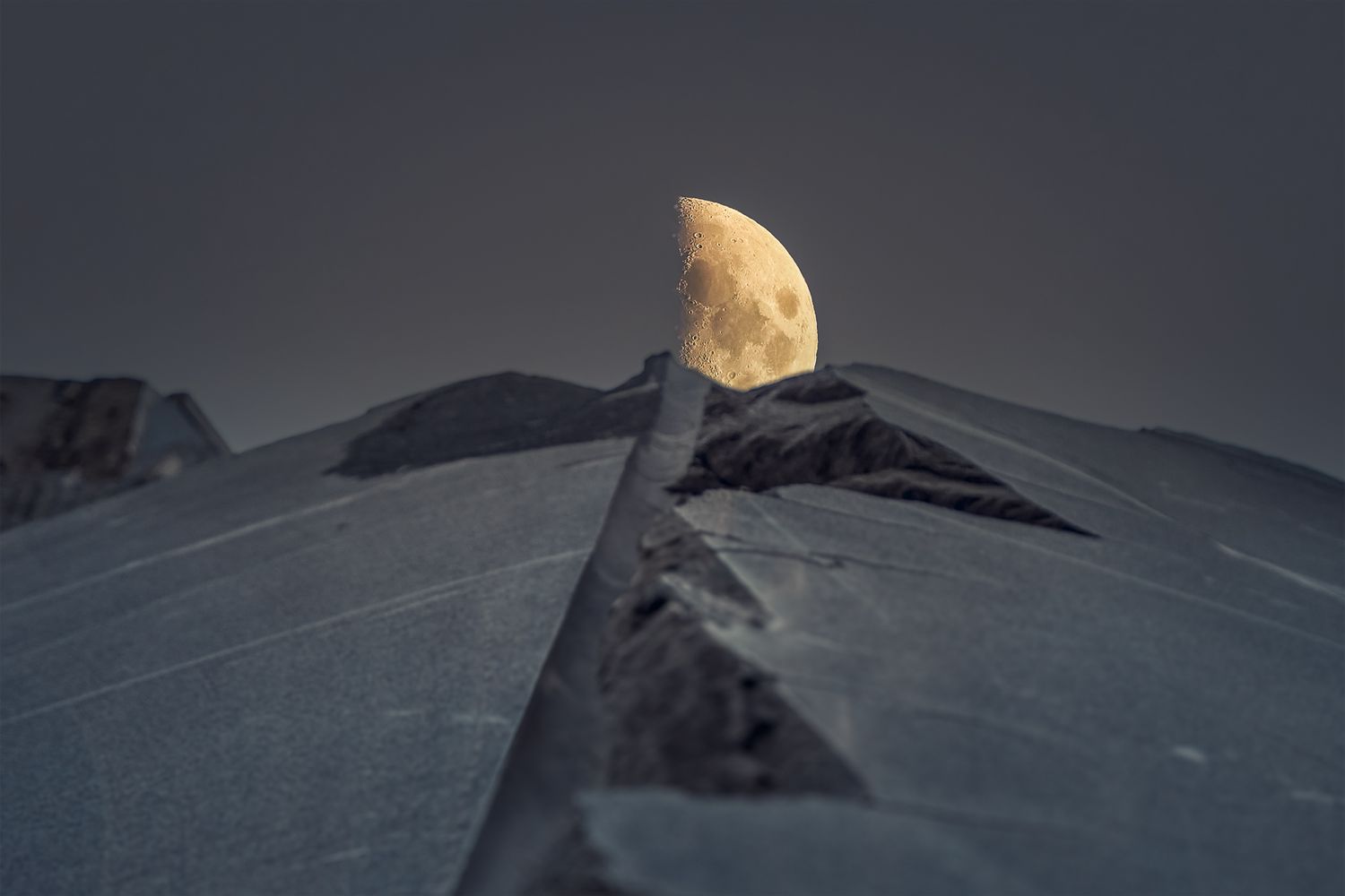 Mezza luna che sorge dietro il profilo frastagliato di una cava di notte; immagine poetica che unisce materia, paesaggio e visione autoriale