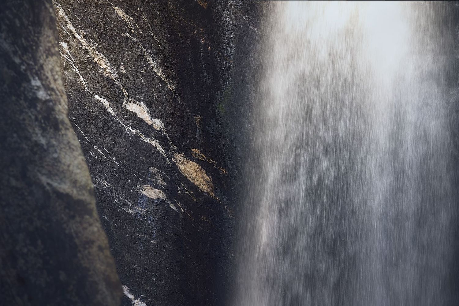 Cascata che scorre tra rocce con parete a sinistra, acqua in movimento e luce morbida che crea un’atmosfera naturale, fresca e contemplativa