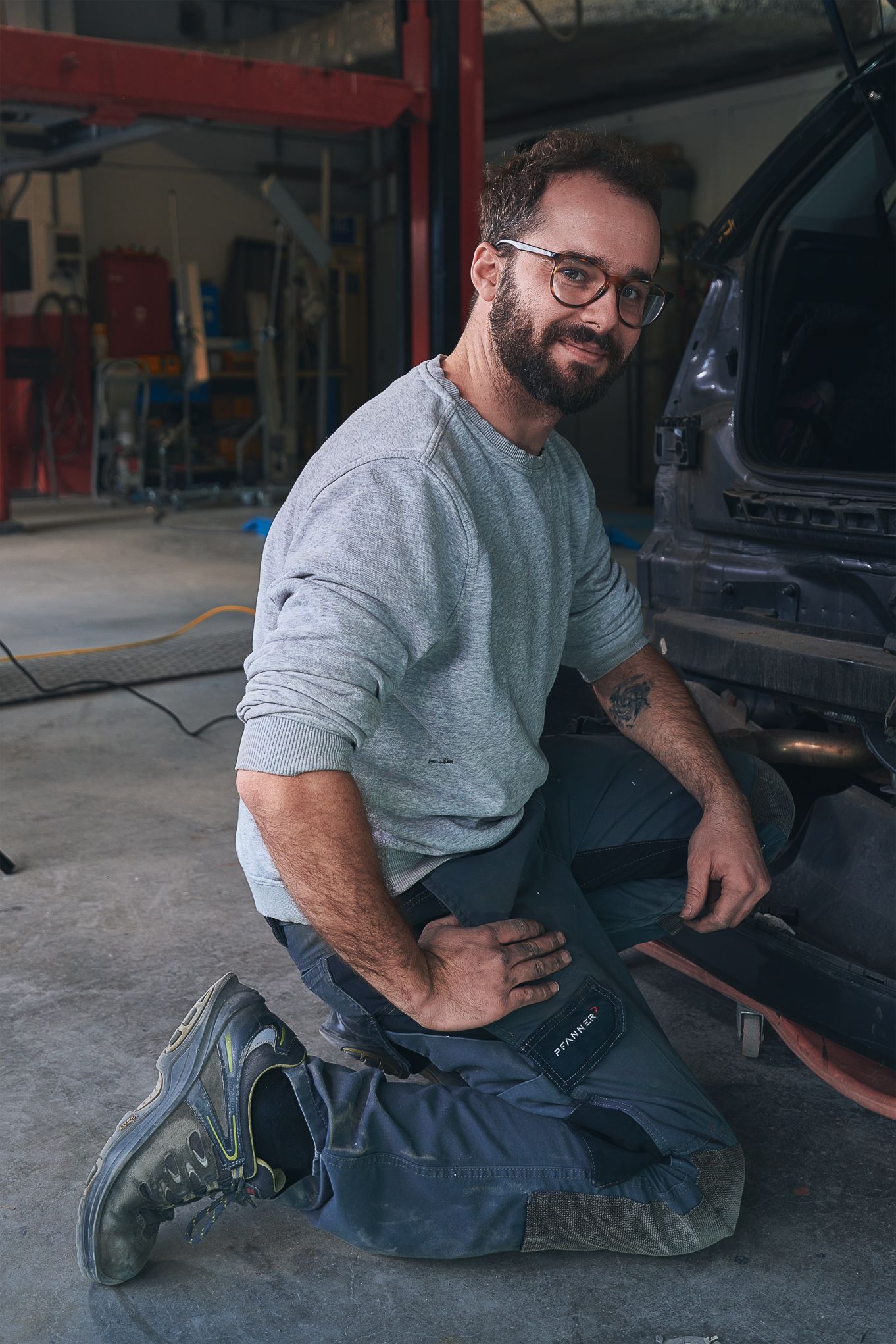 «Ritratto candido di un uomo inginocchiato in officina, con sorriso naturale e luce morbida, colto durante il lavoro in carrozzeria.
