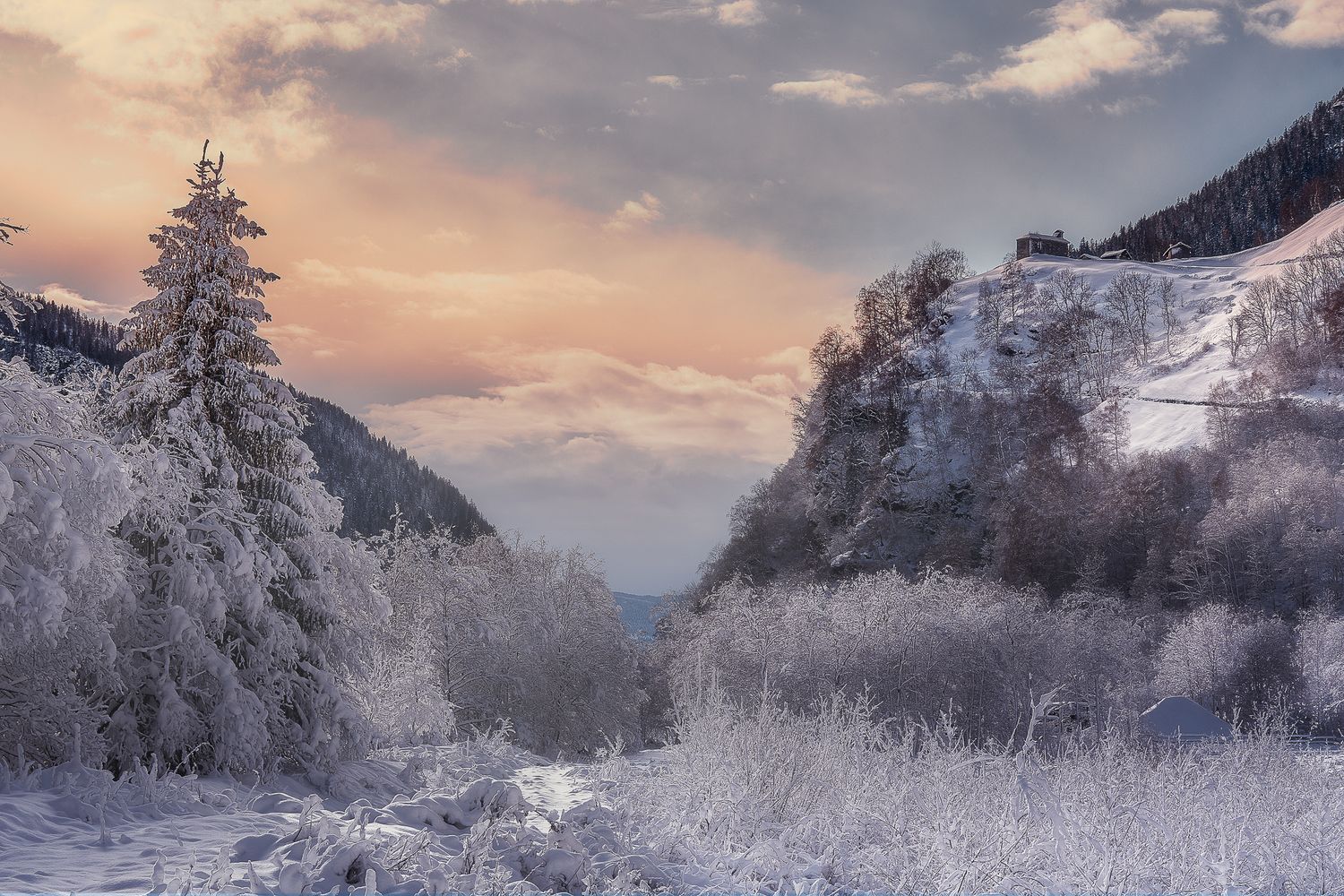 Valle alpina invernale con alberi ghiacciati, crinale innevato e cielo arancione-azzurro al tramonto; piccola costruzione emerge dalla monta