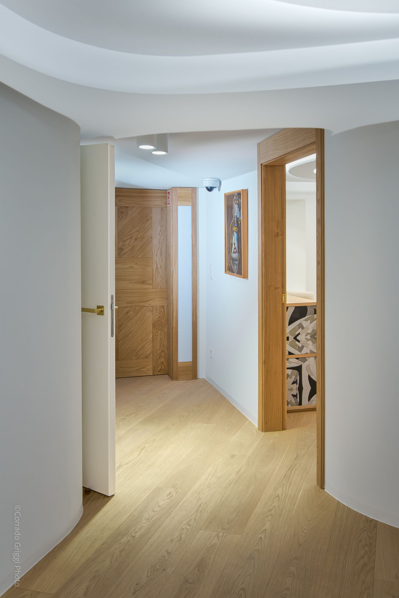 Corridoio moderno con pareti curve, porte in legno e illuminazione soffusa che conduce a diversi ambienti, creando un’atmosfera ordinata.