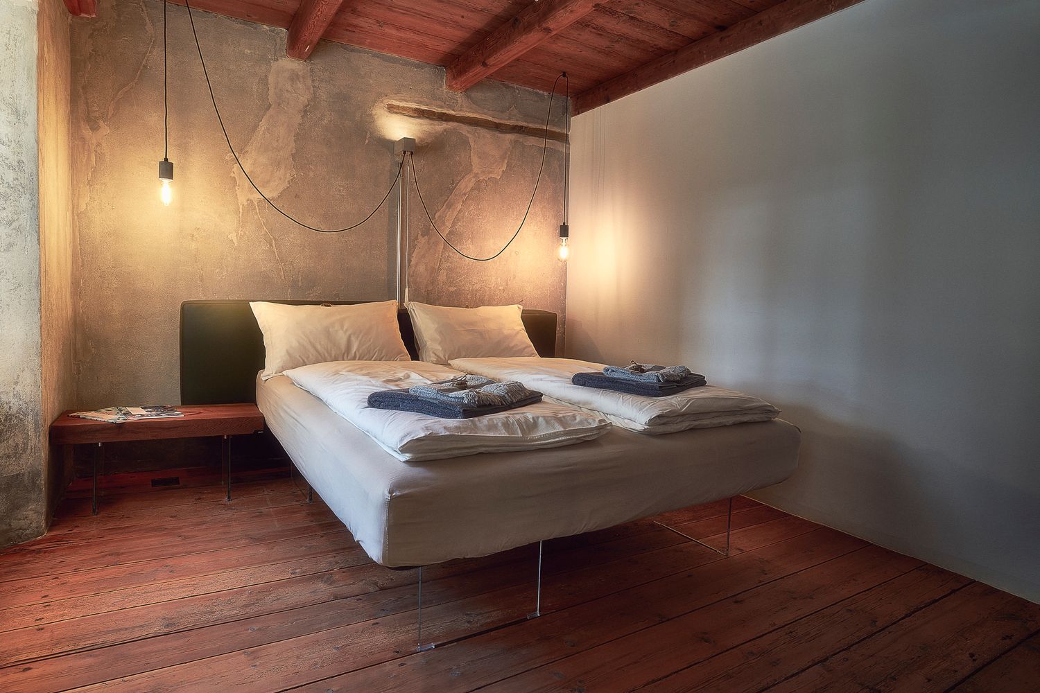 Camera da letto con pareti materiche, soffitto in legno e letto sospeso, illuminata da luci pendenti che creano un’atmosfera calda.