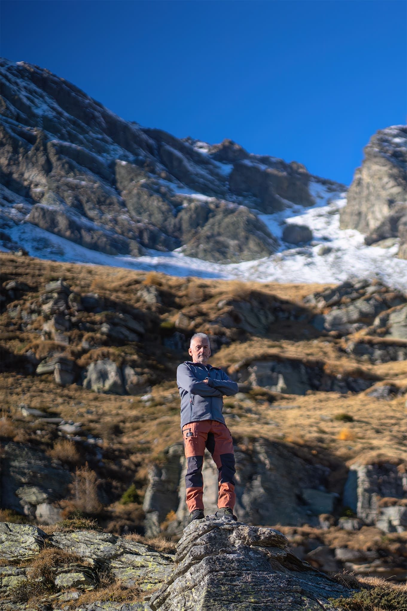 Fotografo in piedi su uno spuntone di roccia, una vasta montagna sullo sfondo; ritratto ambientato che unisce presenza, paesaggio e sguardo
