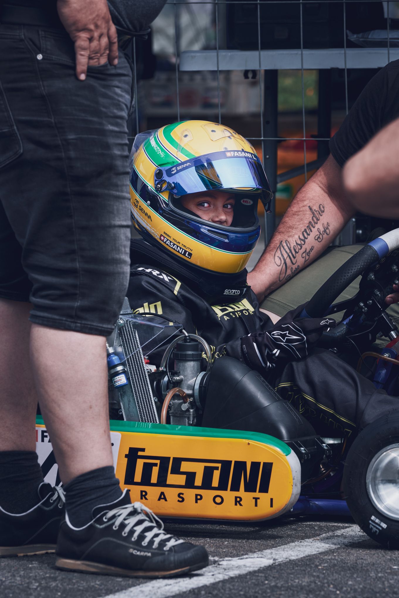 «Giovane pilota di go-kart con tuta e casco, circondato da mani che lo rassicurano prima della gara, occhi che sorridono dietro la visiera.»