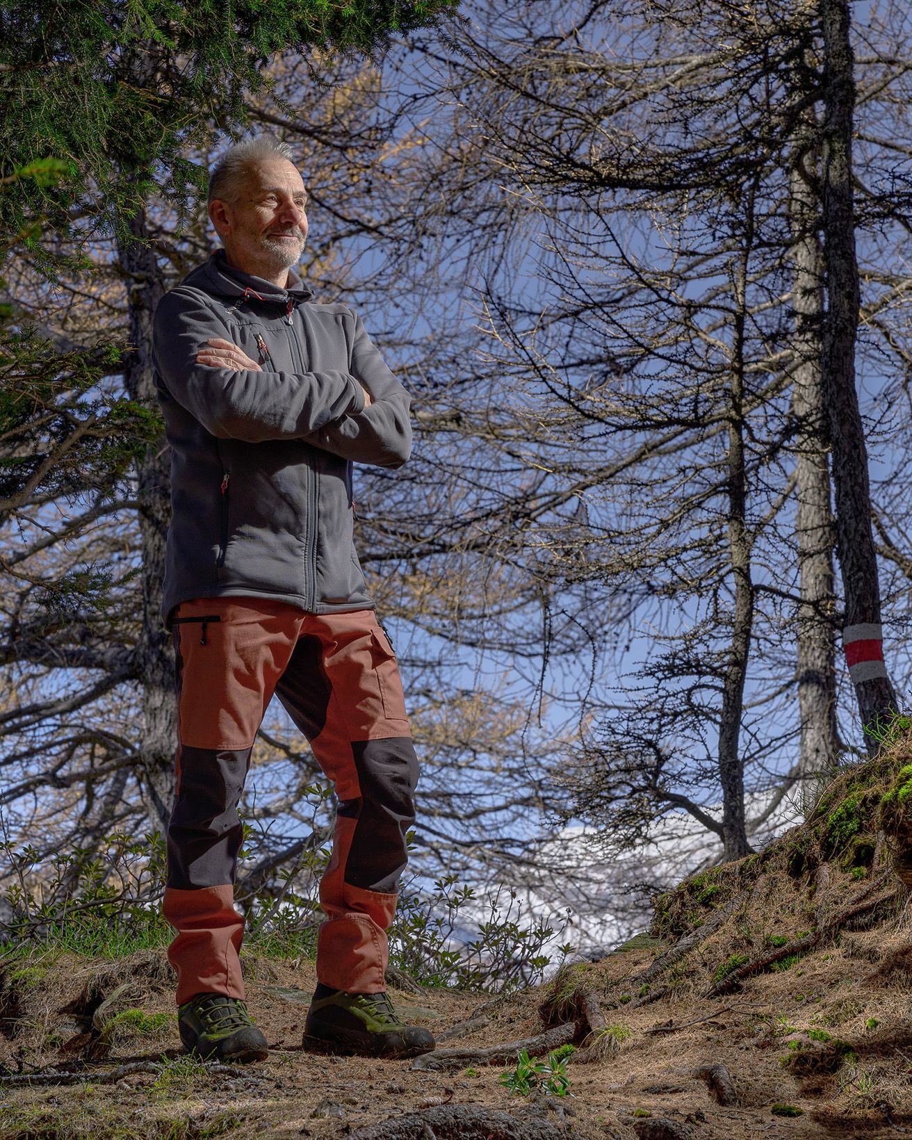 Ritratto ambientato di fotografo in contesto montano con bosco di larici e abeti sullo sfondo.