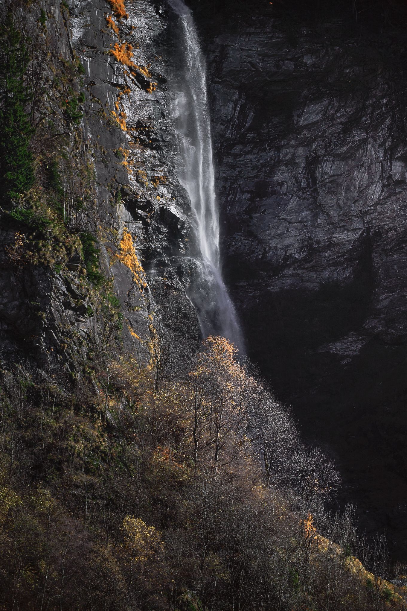 Cascata alpina che scorre tra pareti di roccia scura, dettagli di venature minerali e acqua in movimento morbido.