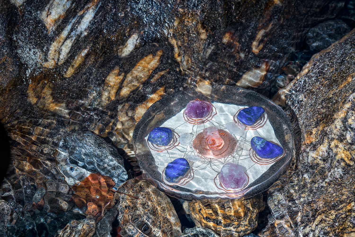 Armonizzatore artigianale immerso in acqua trasparente, circondato da rifrazioni luminose su roccia dorata: materia, luce e movimento