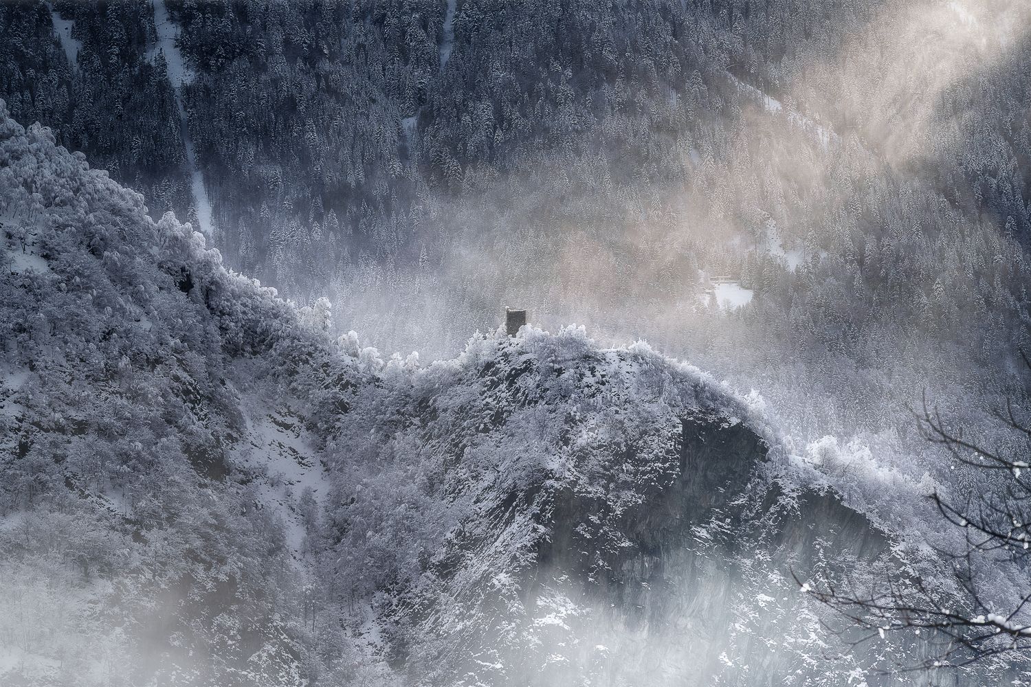 Antica torre su crinale alpino innevato avvolto da nebbia e luce filtrante, paesaggio invernale suggestivo e remoto.