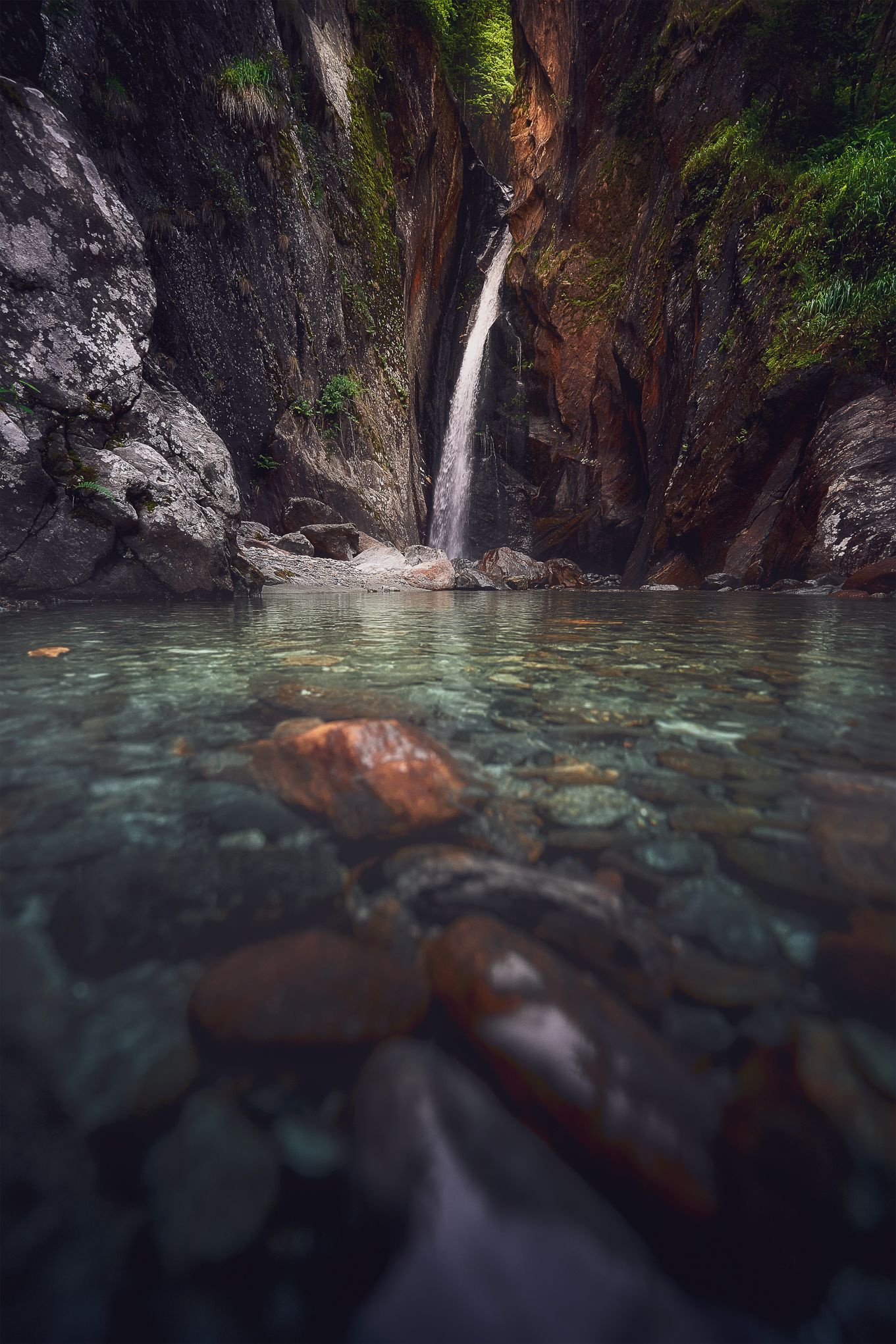 Cascata in gola di roccia con laghetto turchese e massi in primo piano, luce soffusa tra le pareti e atmosfera selvaggia.