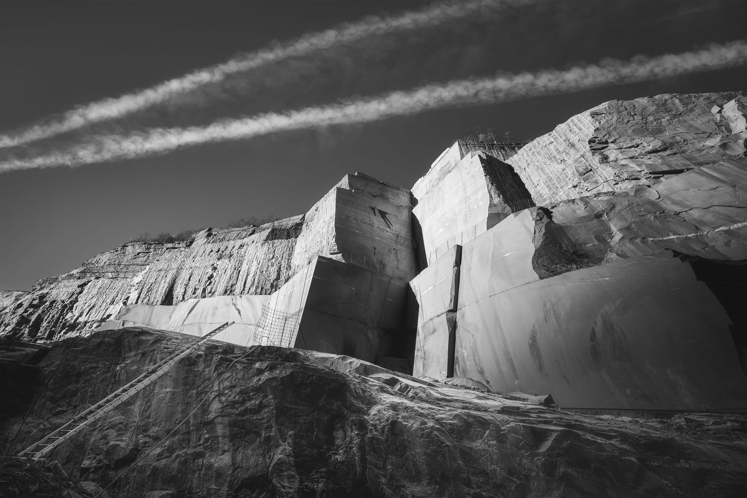 Pareti e blocchi monumentali nella cava Polti ad Arvigo (Grigioni), ripresa dal basso con scala e cielo attraversato da scie di aereo.