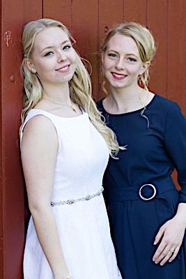 Two people in white and navy dresses pose together against a red wooden wall backdrop.