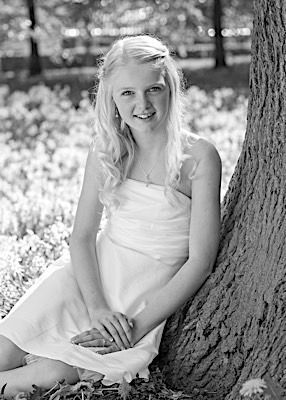 Black and white portrait of person in white dress sitting by tree in sunlit garden.