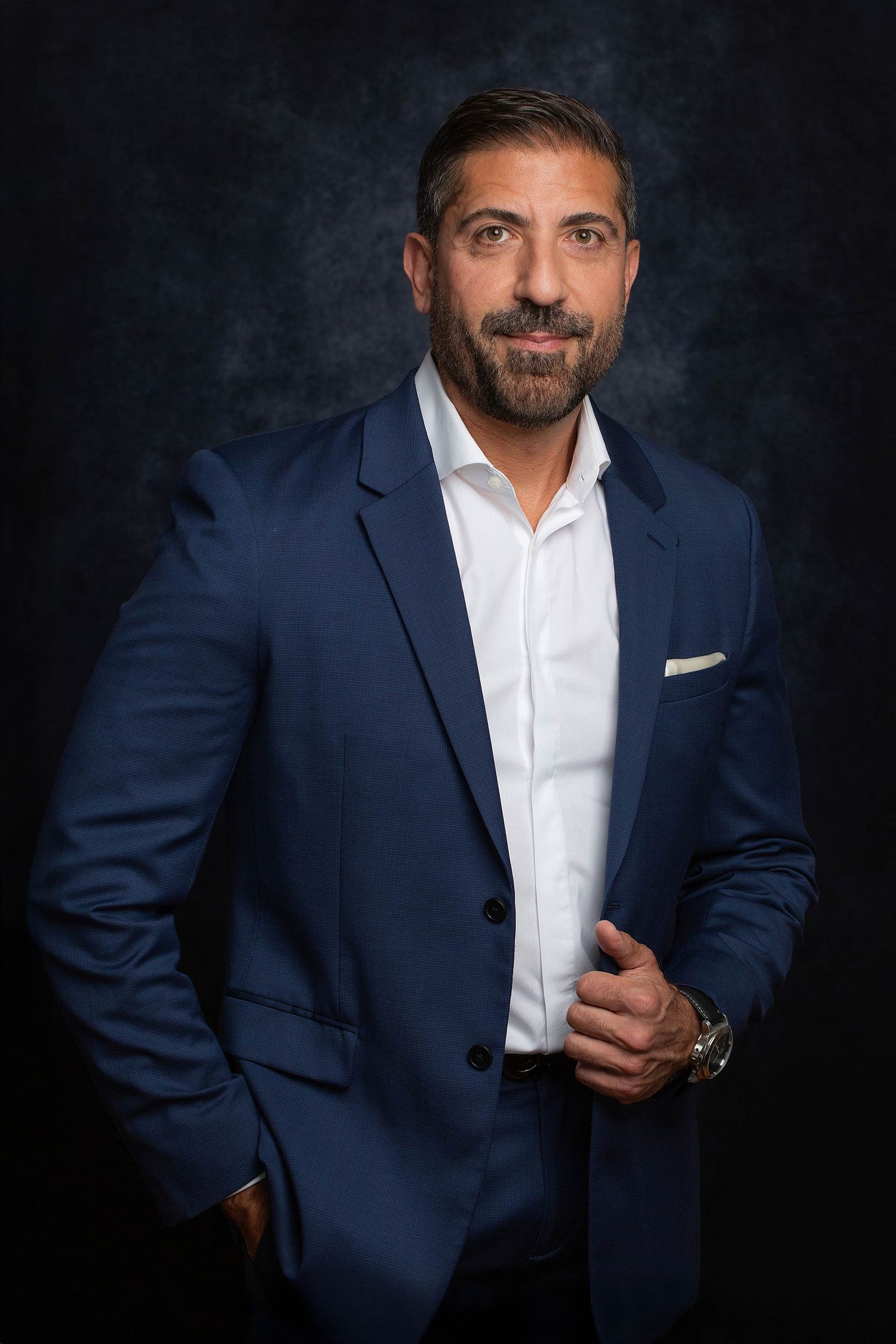 Business portrait in navy suit and white shirt against dark backdrop showing confident pose.