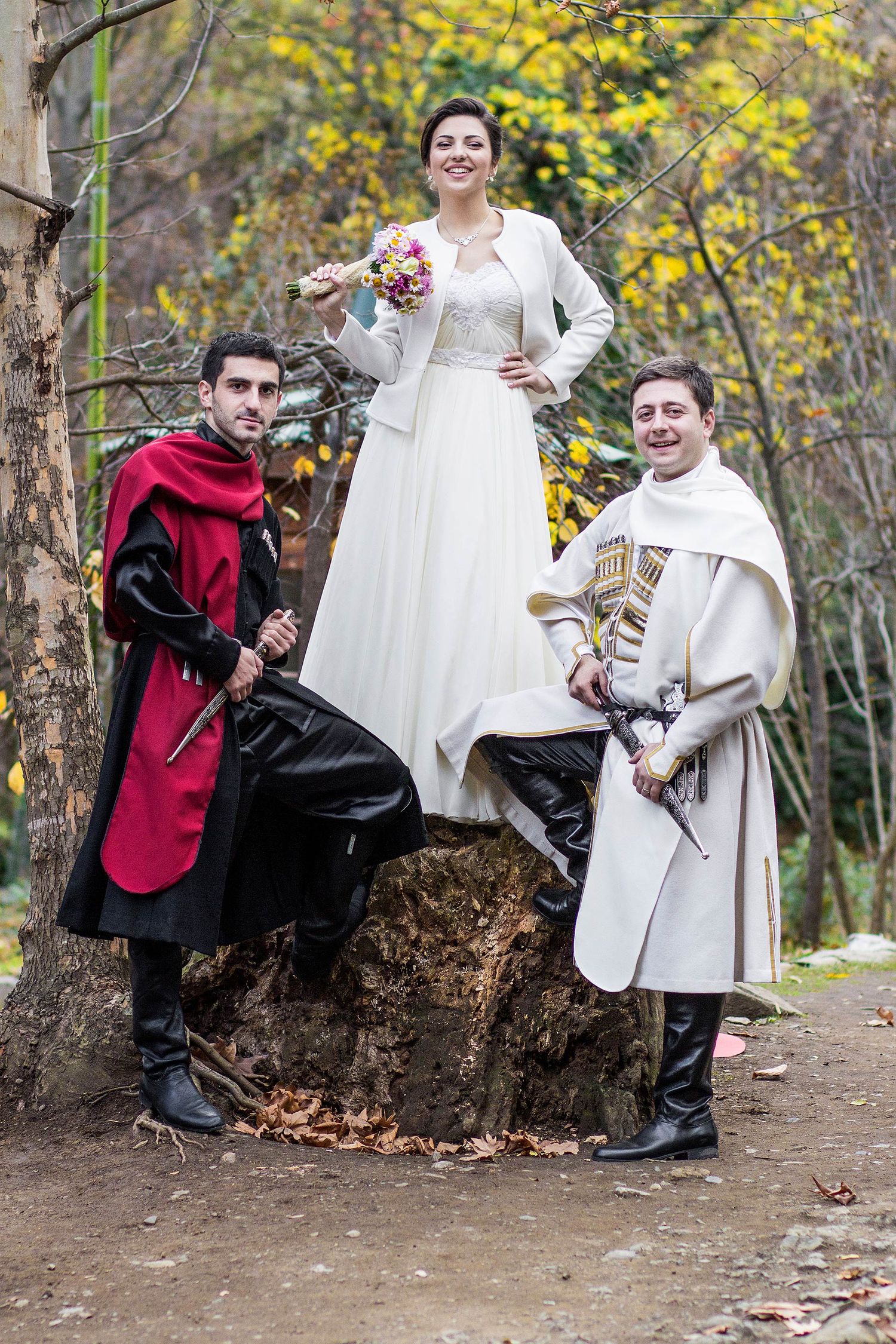 Three people in medieval costumes pose playfully on a fallen tree trunk in an autumn forest.