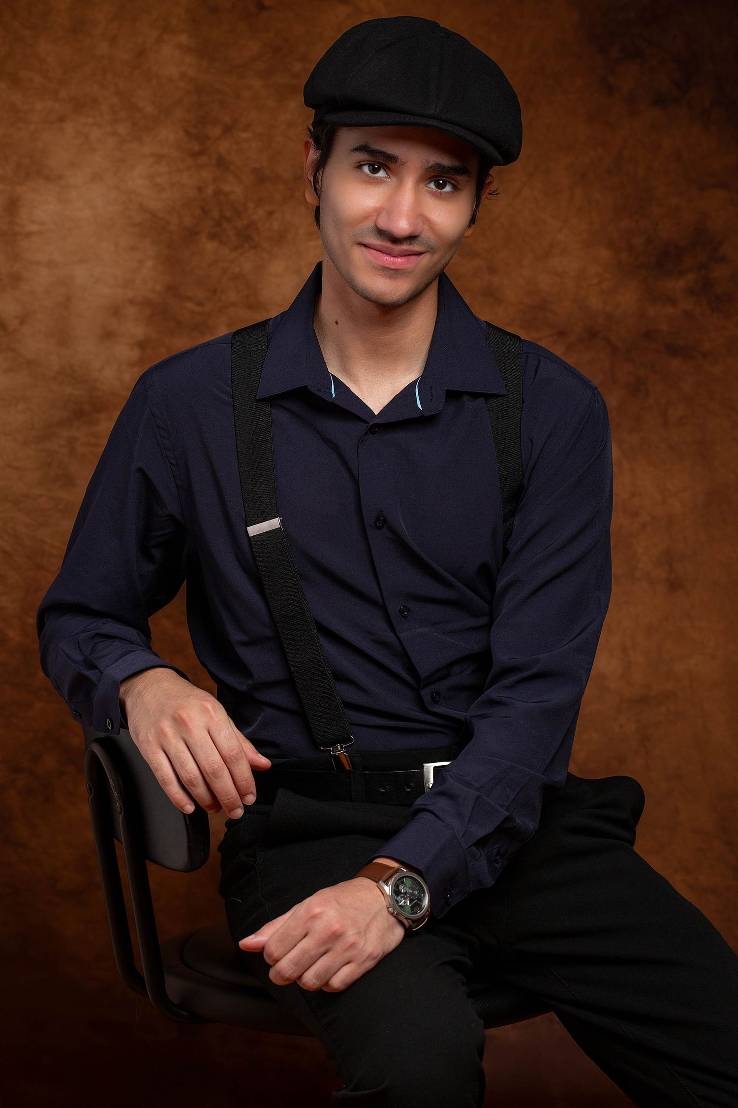 Figure in dark outfit and cap poses in studio against brown backdrop.