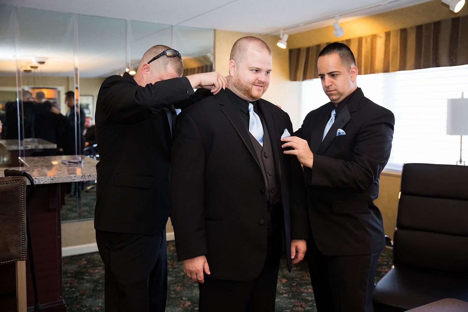 Groomsmen helping adjust formal attire in a hotel room before the wedding ceremony.