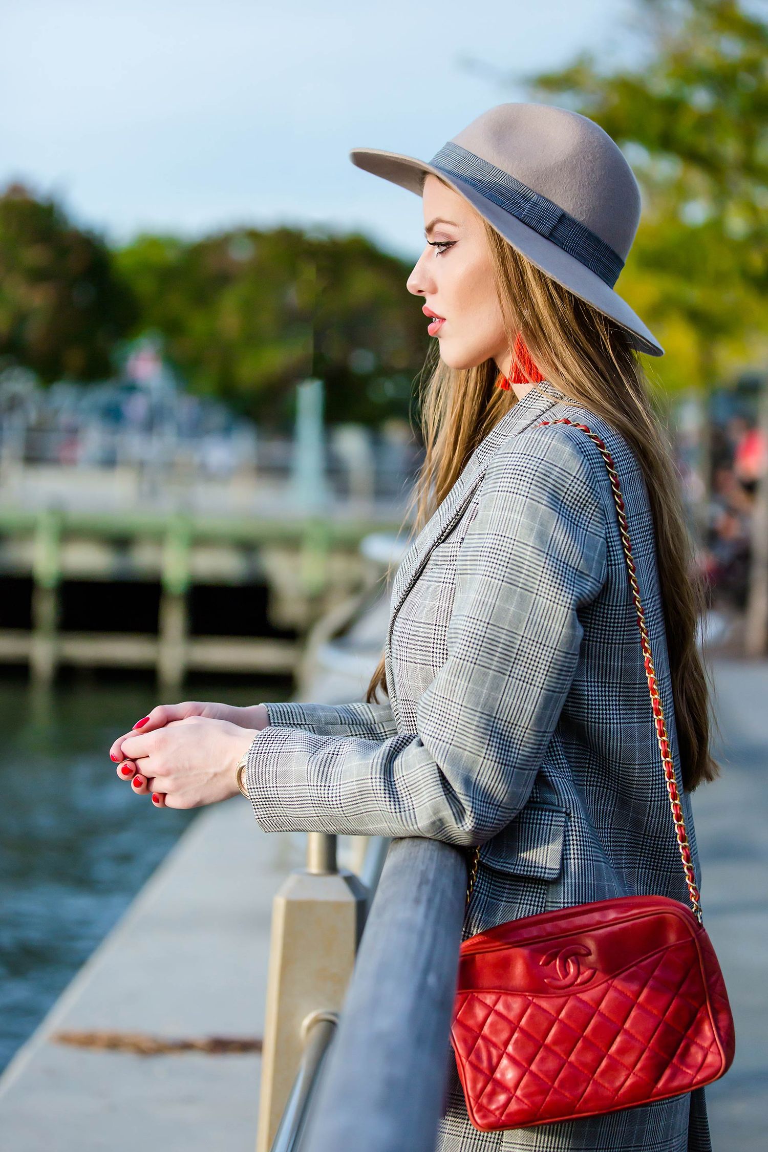 Grey fedora and plaid blazer outfit with red quilted bag by waterfront.