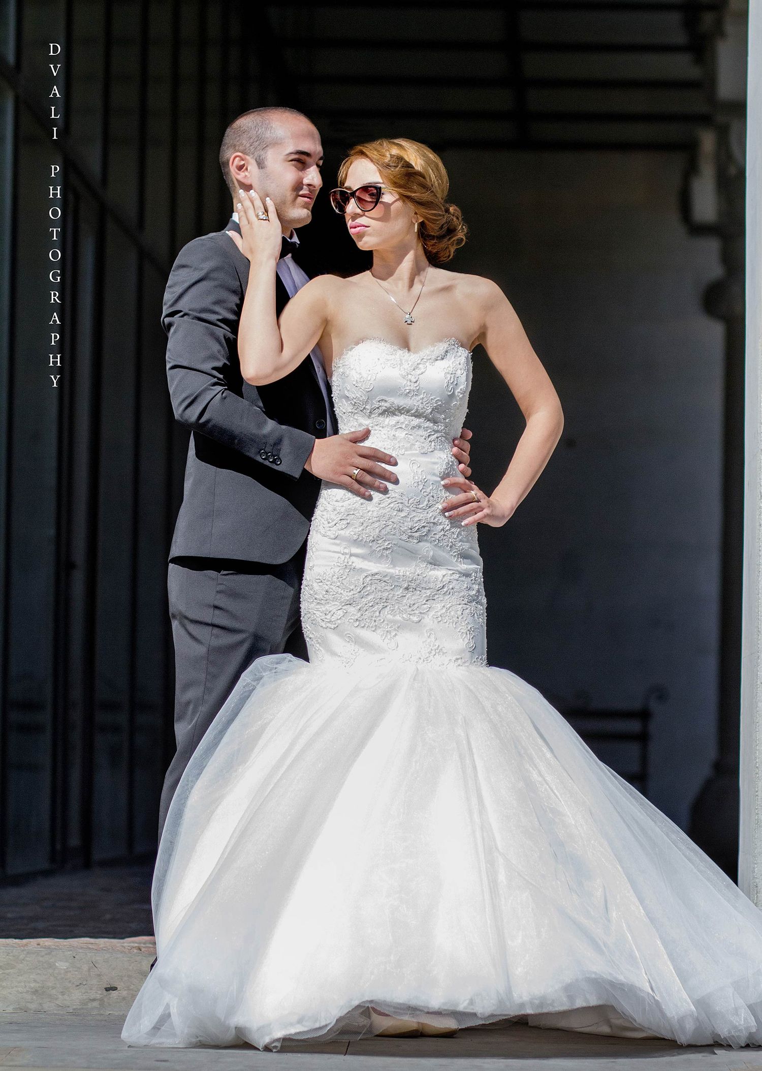 A stylish couple poses dramatically in formal wedding attire against a dark background.