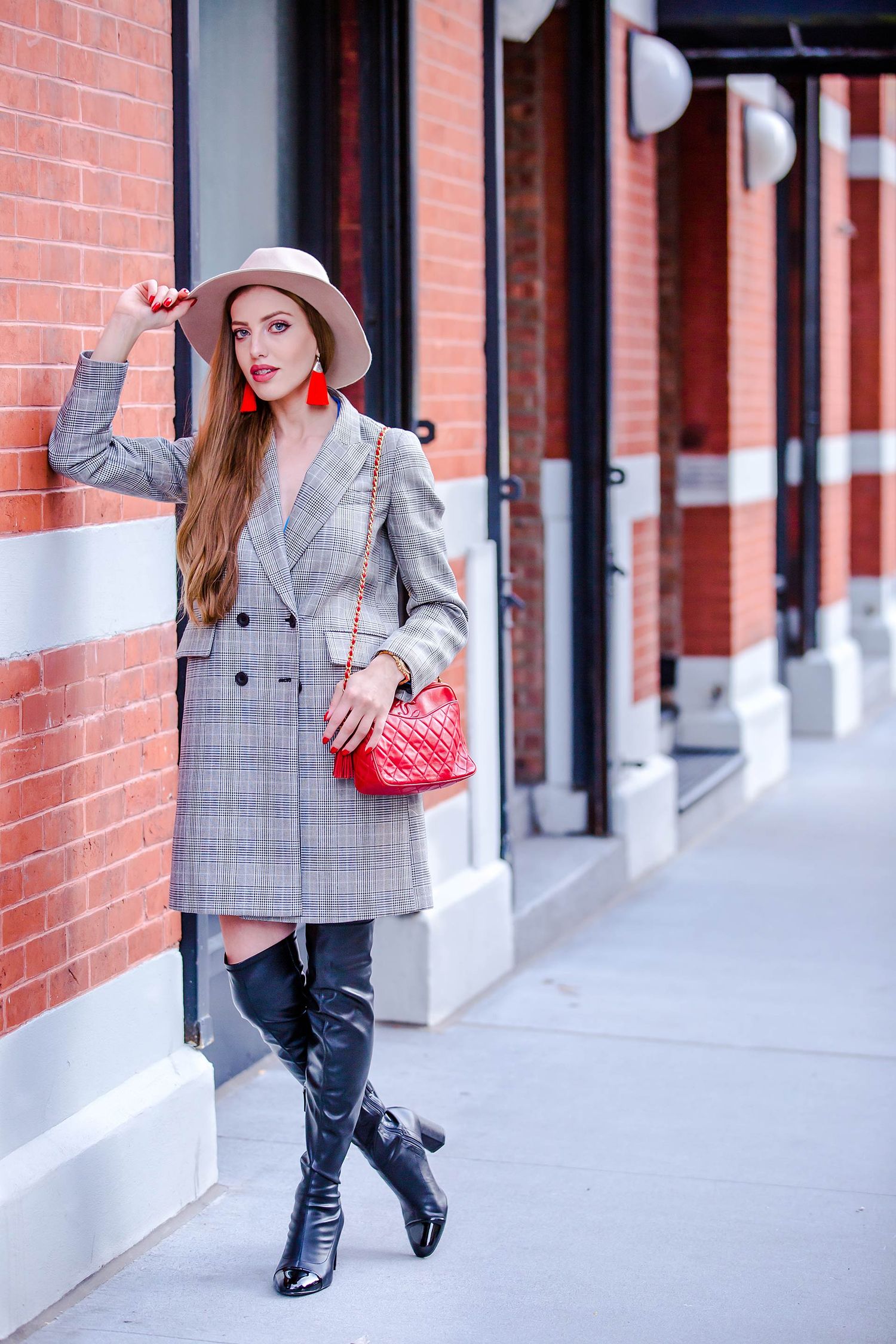 Woman in gray plaid blazer and red hat posing against brick building wall.