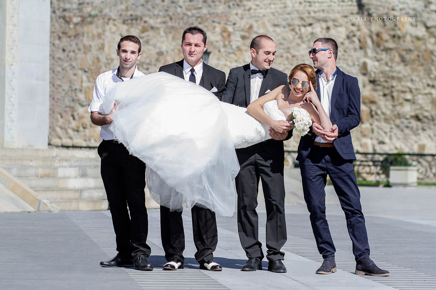 Wedding party members playfully carry a bride in her flowing white dress outdoors.