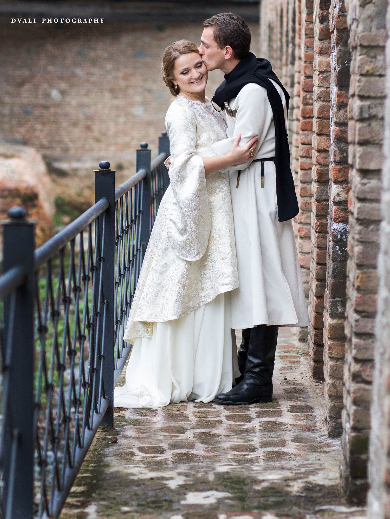 A romantic medieval-inspired couple in white embraces along a brick wall and iron railing.