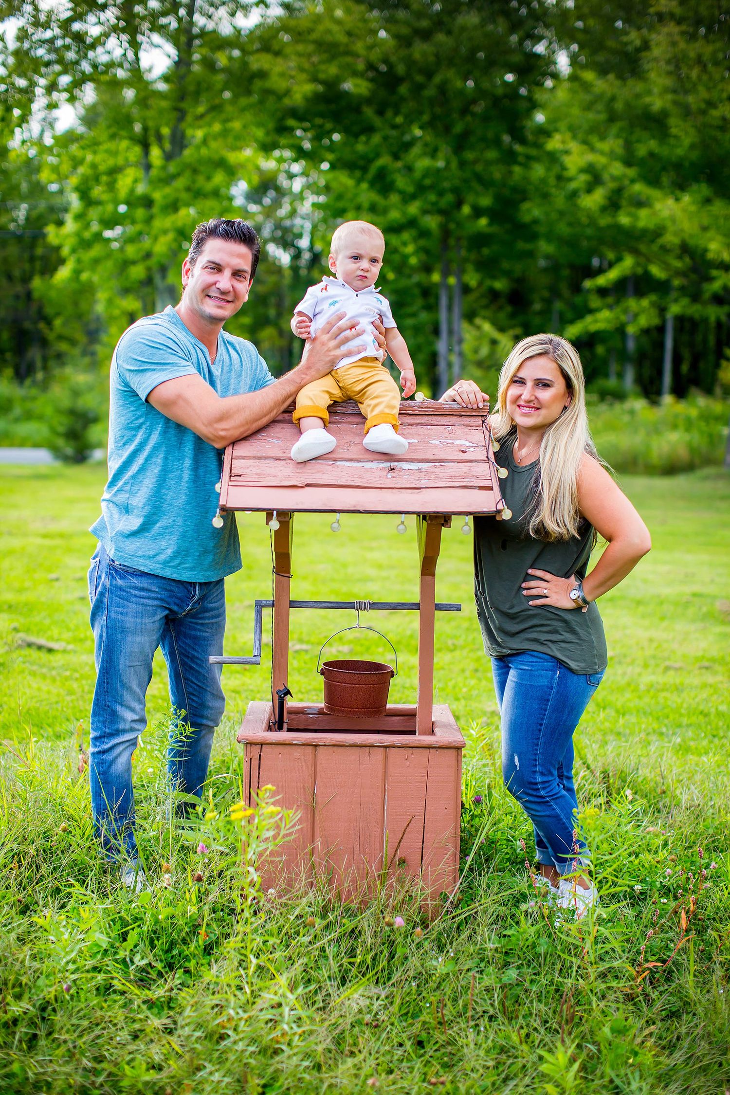 Playful couple posing at rustic wooden well structure in green meadow.