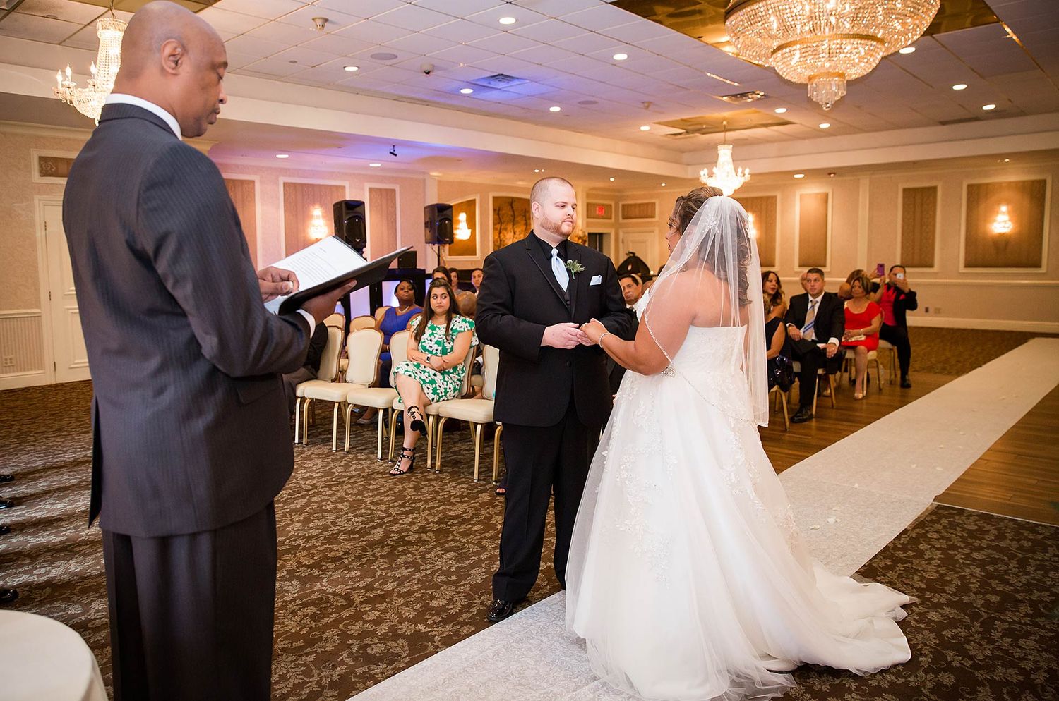 A wedding ceremony takes place in an elegant ballroom with crystal chandeliers and carpeted floors.