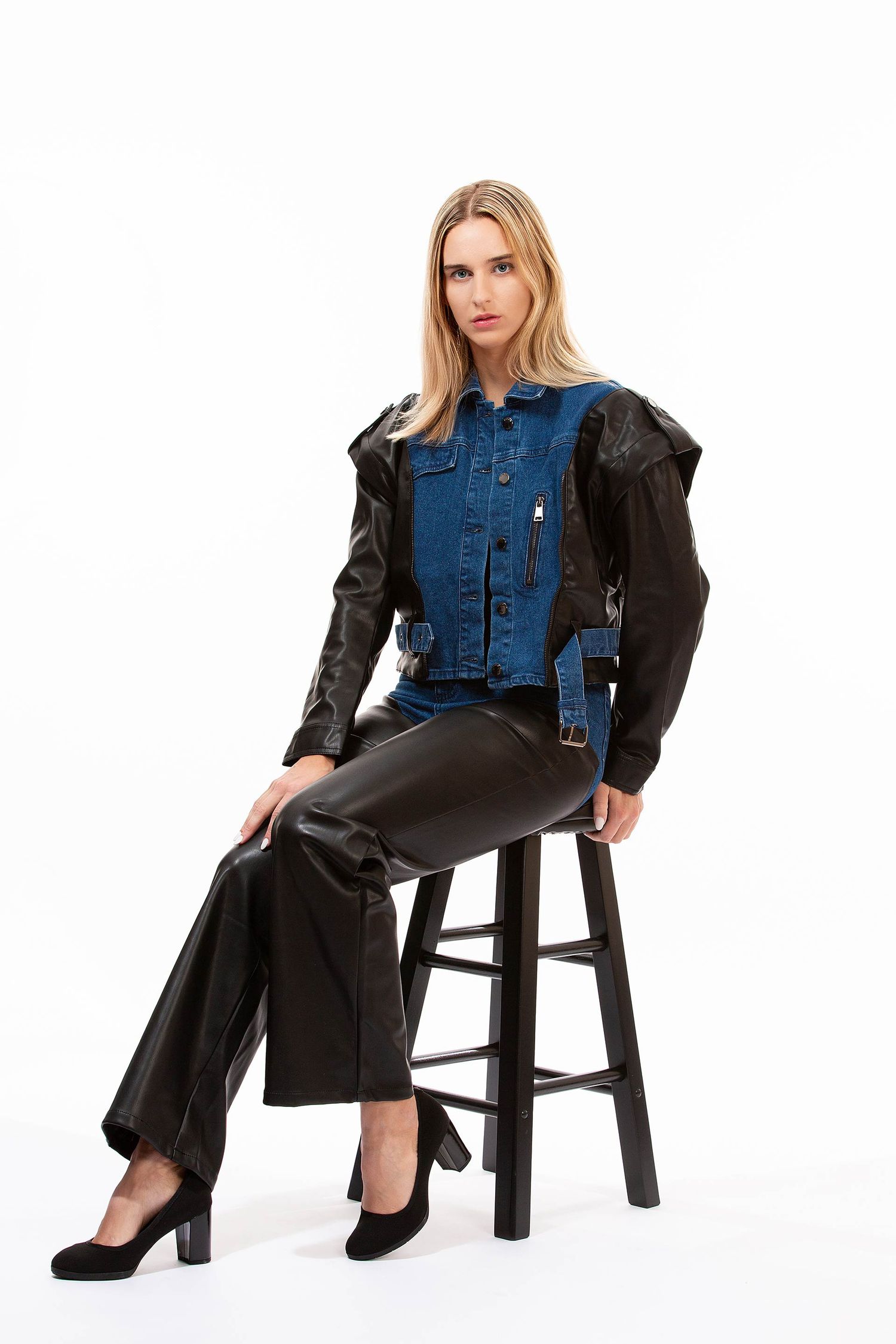 Model seated on wooden stool wearing denim jacket and leather pants in studio setting.