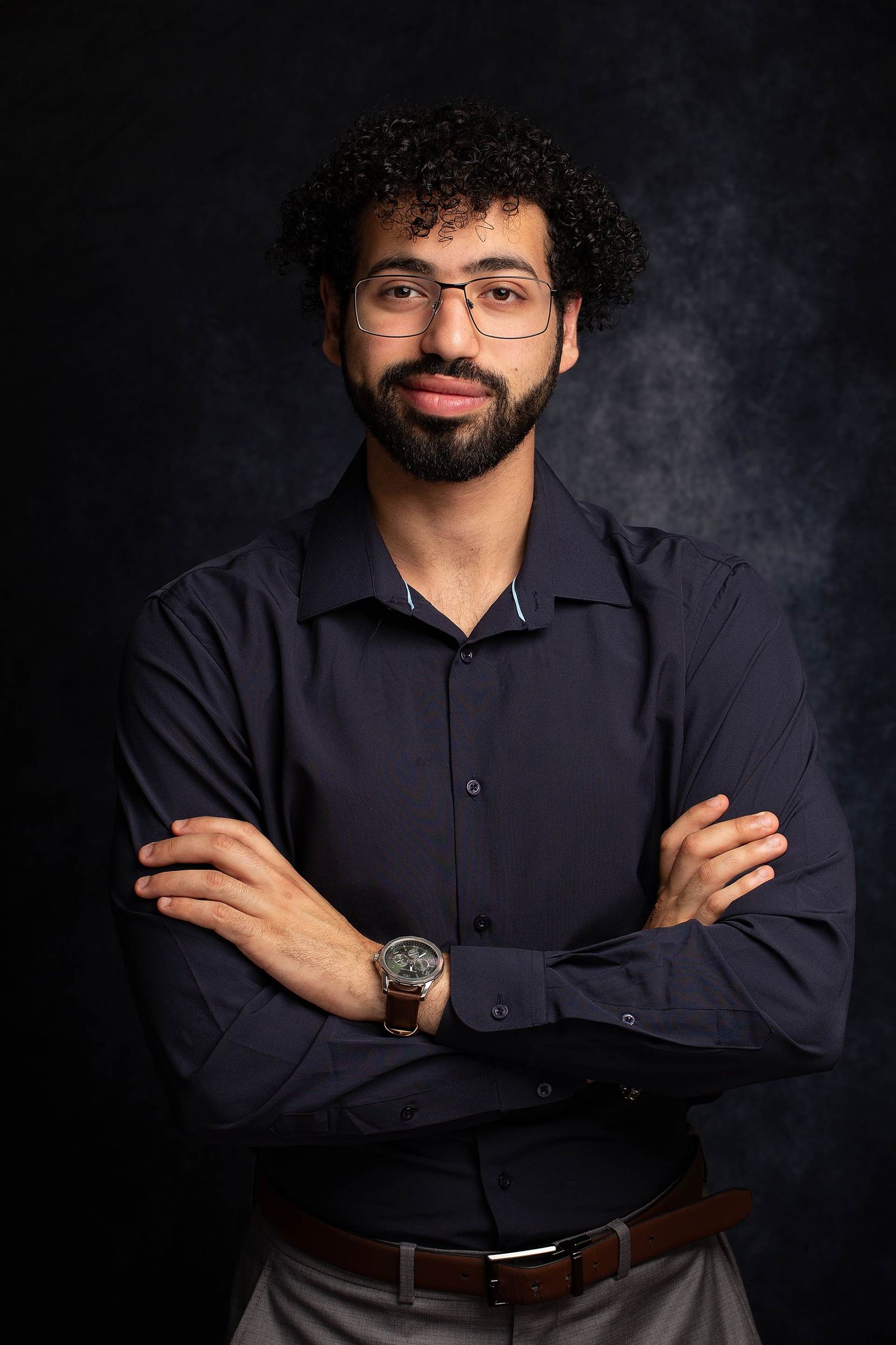 Professional portrait of person in dark blue button-down shirt with crossed arms against gray backdrop.