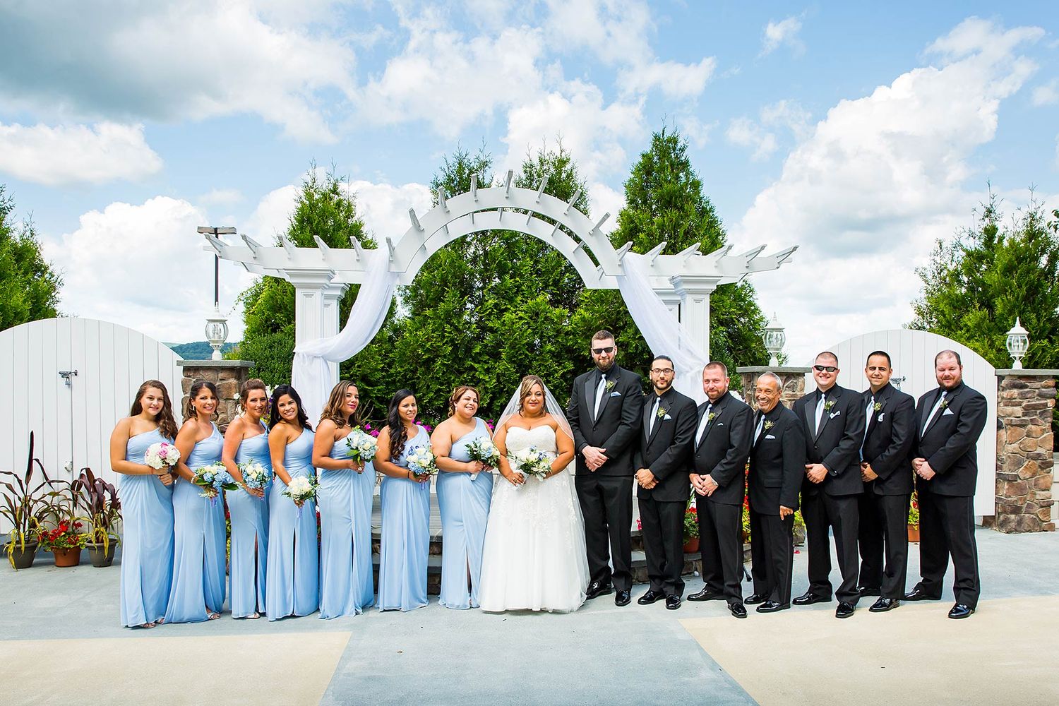 Wedding party in light blue and black formal attire posed under white garden arch outdoors.