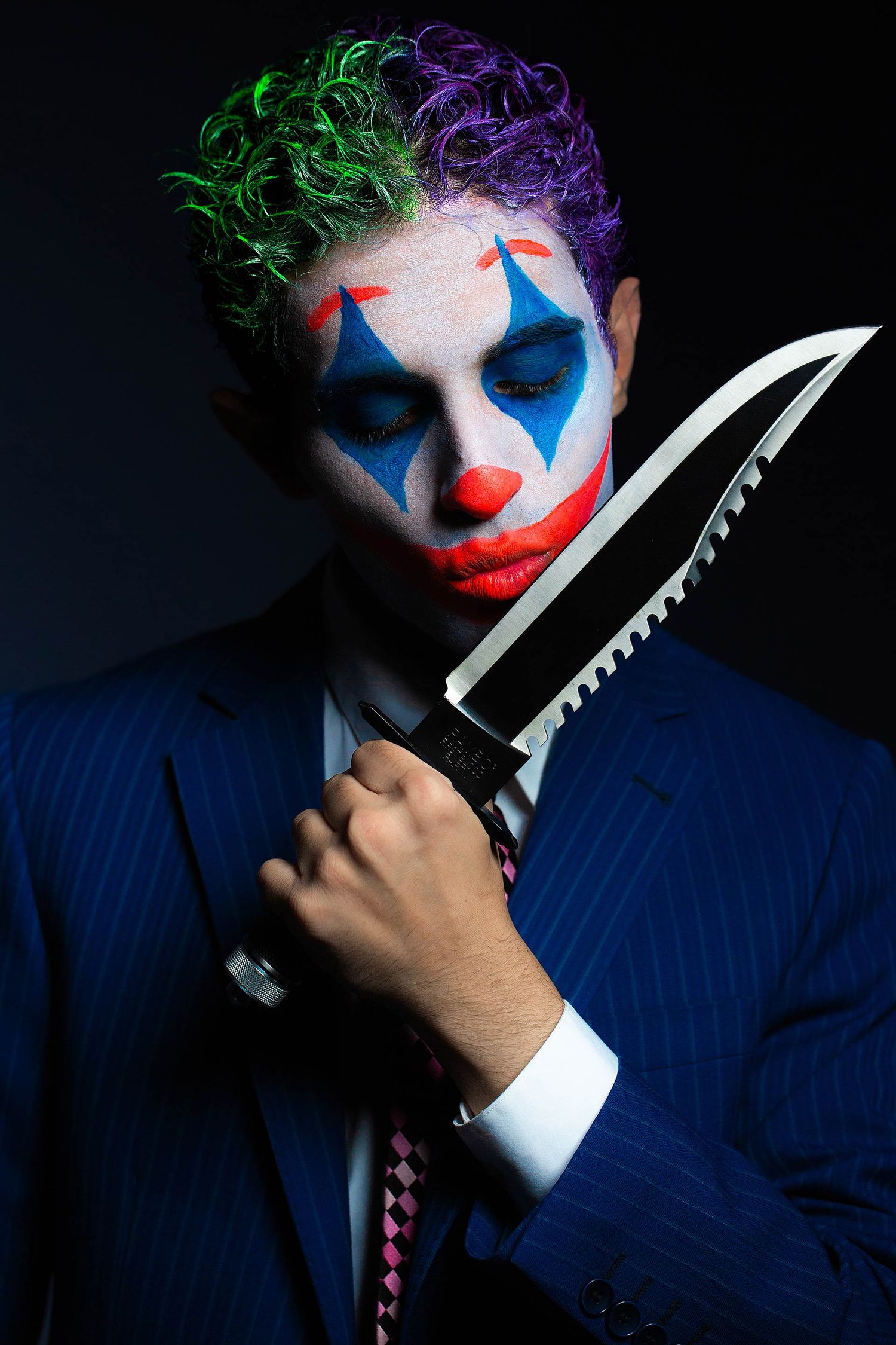 Dramatic clown makeup portrait with blue suit and knife against dark background.
