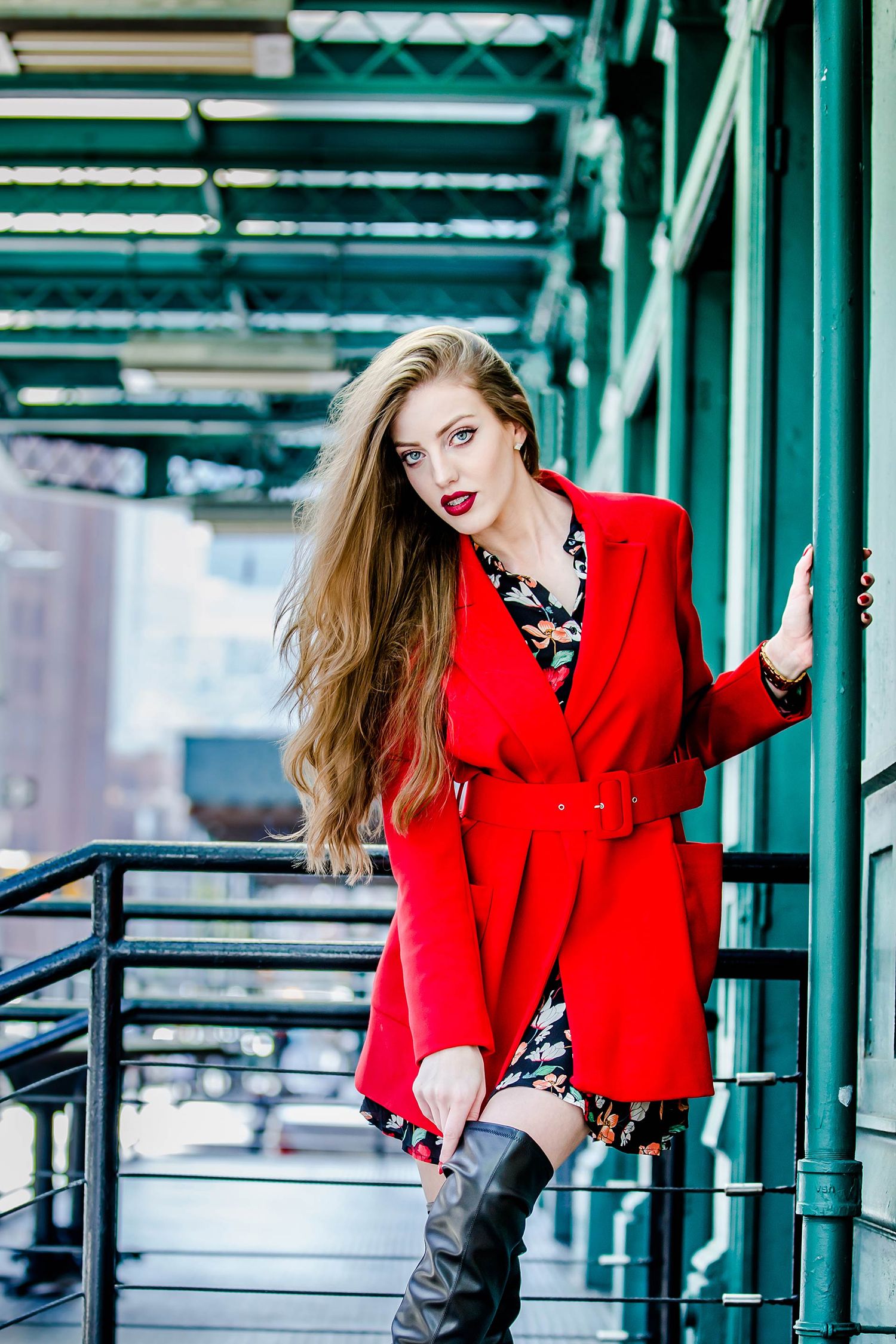Model in striking red coat poses dramatically against industrial green metal architecture.