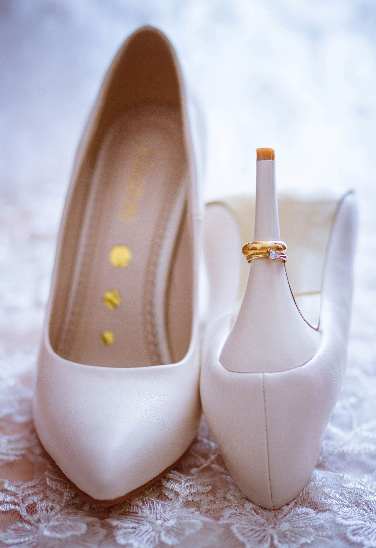 White wedding heels with engagement rings balanced on shoe heel against lace background.