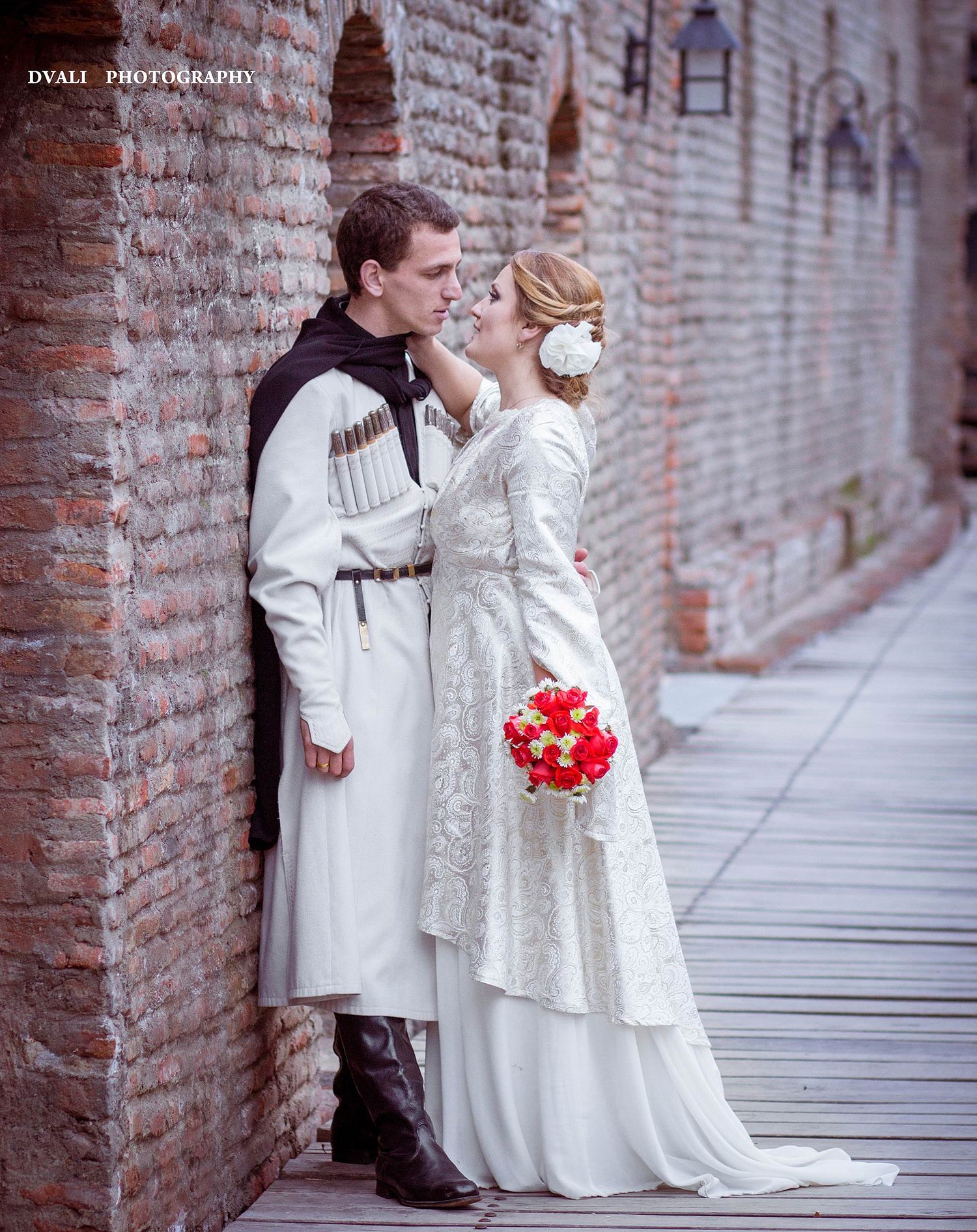 A romantic medieval wedding photo with couple against brick wall holding red bouquet.