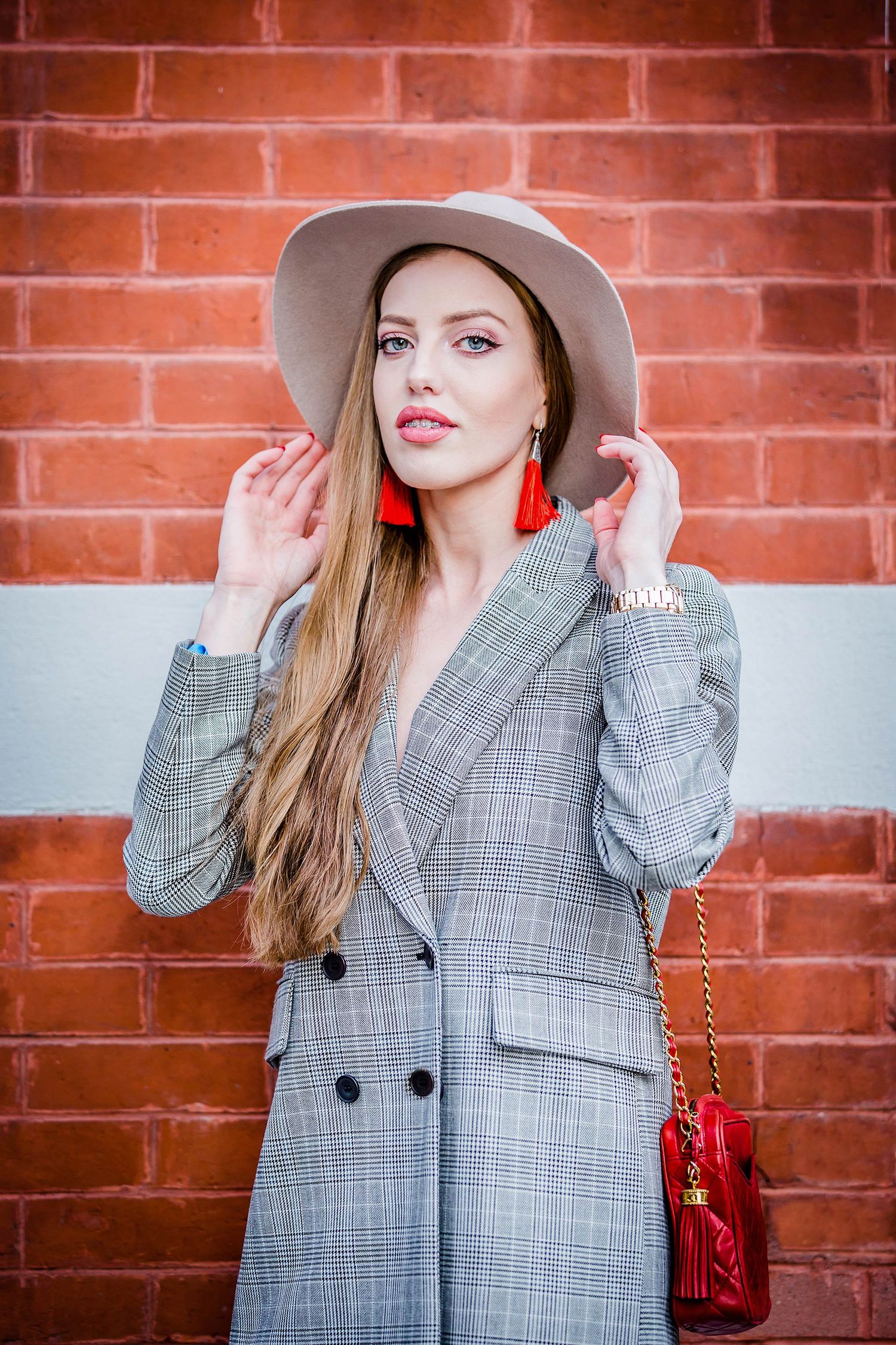 Grey plaid blazer styled with fedora and red statement earrings against brick wall.