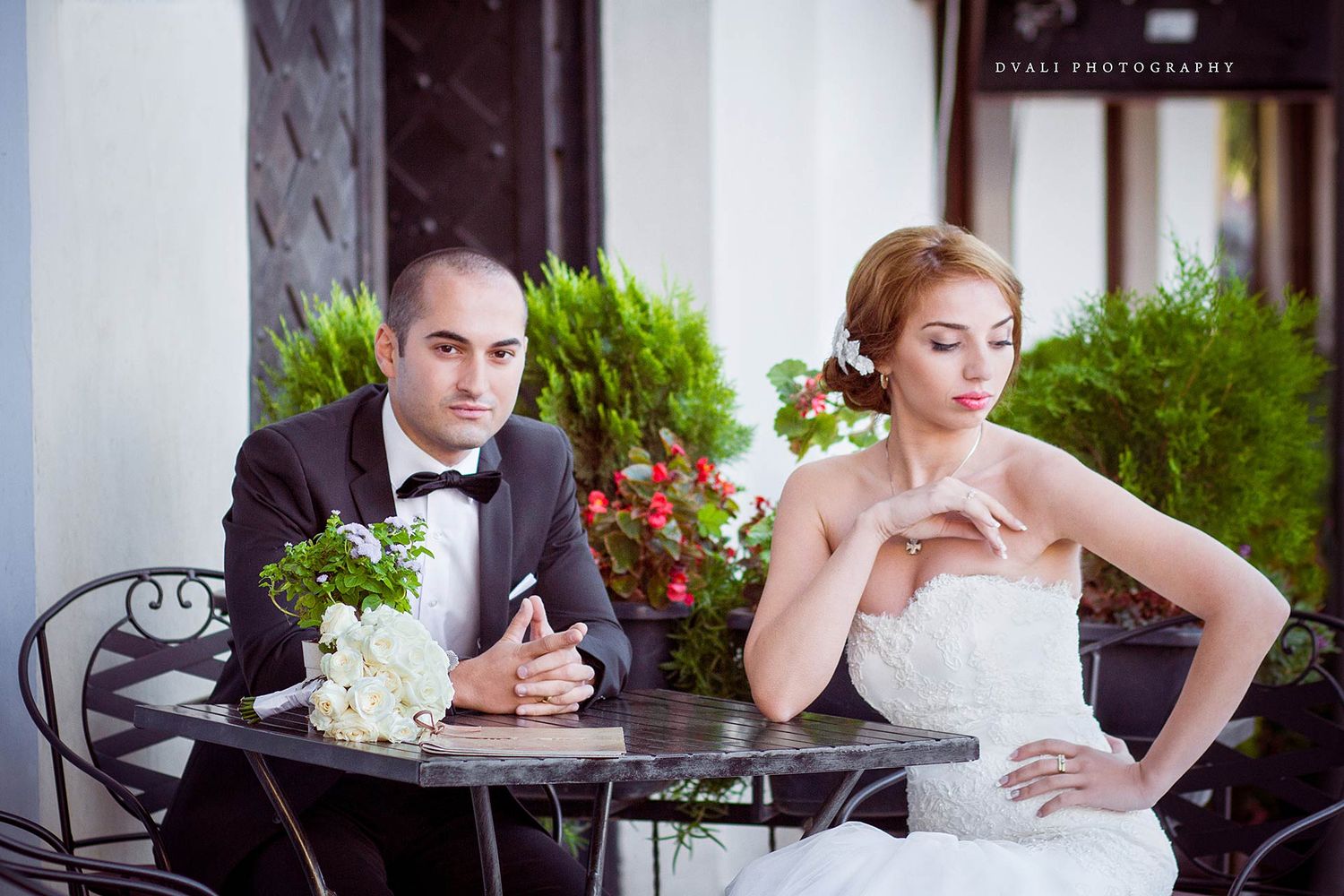 An elegant wedding couple seated at a table outdoors in formal attire.