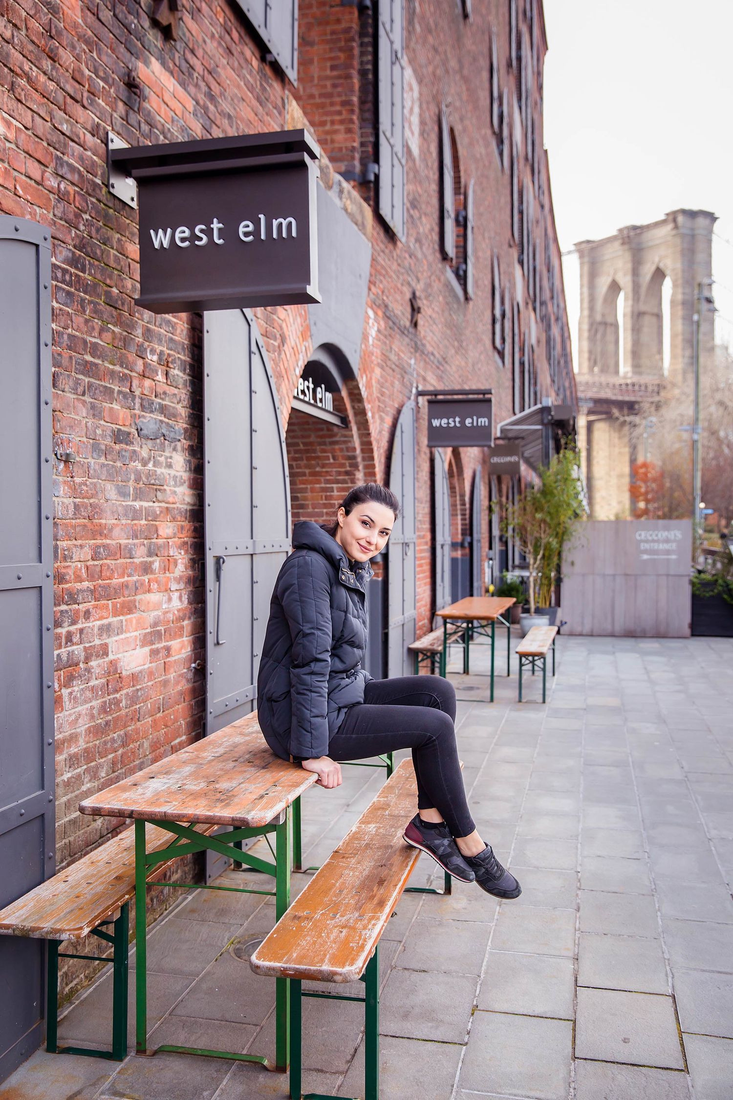 Person sitting on wooden bench outside West Elm store with brick building facade.