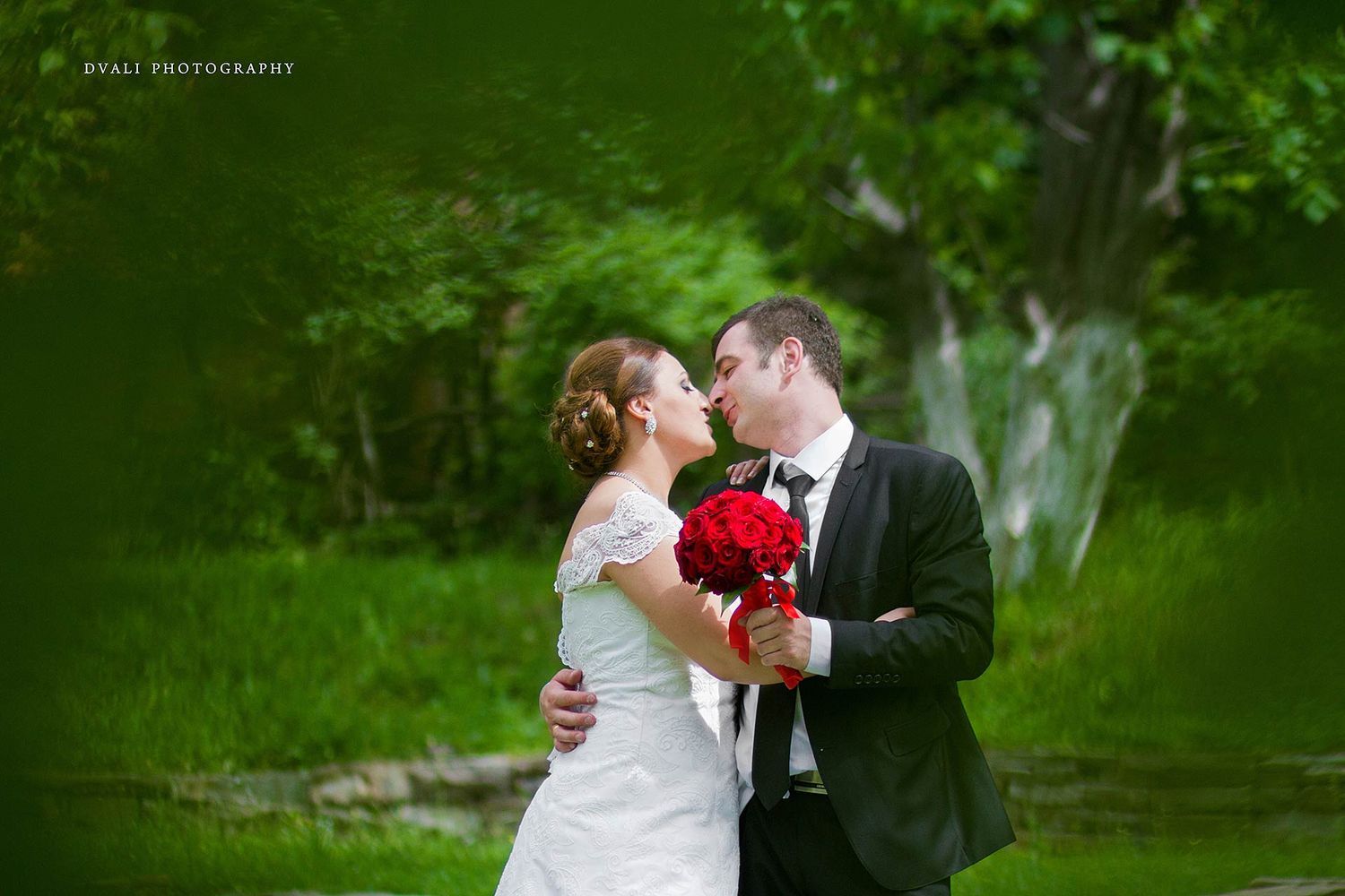 Romantic wedding portrait with red bouquet in lush green outdoor setting.