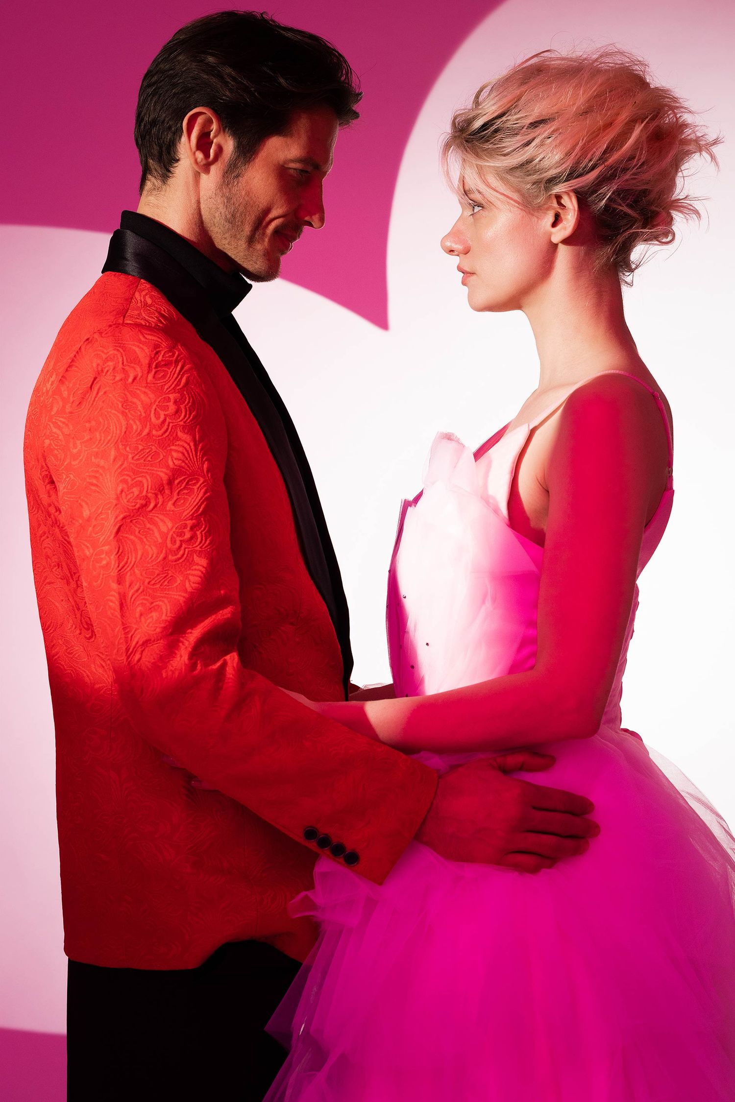 Couple dancing in formal attire against pink and white backdrop.